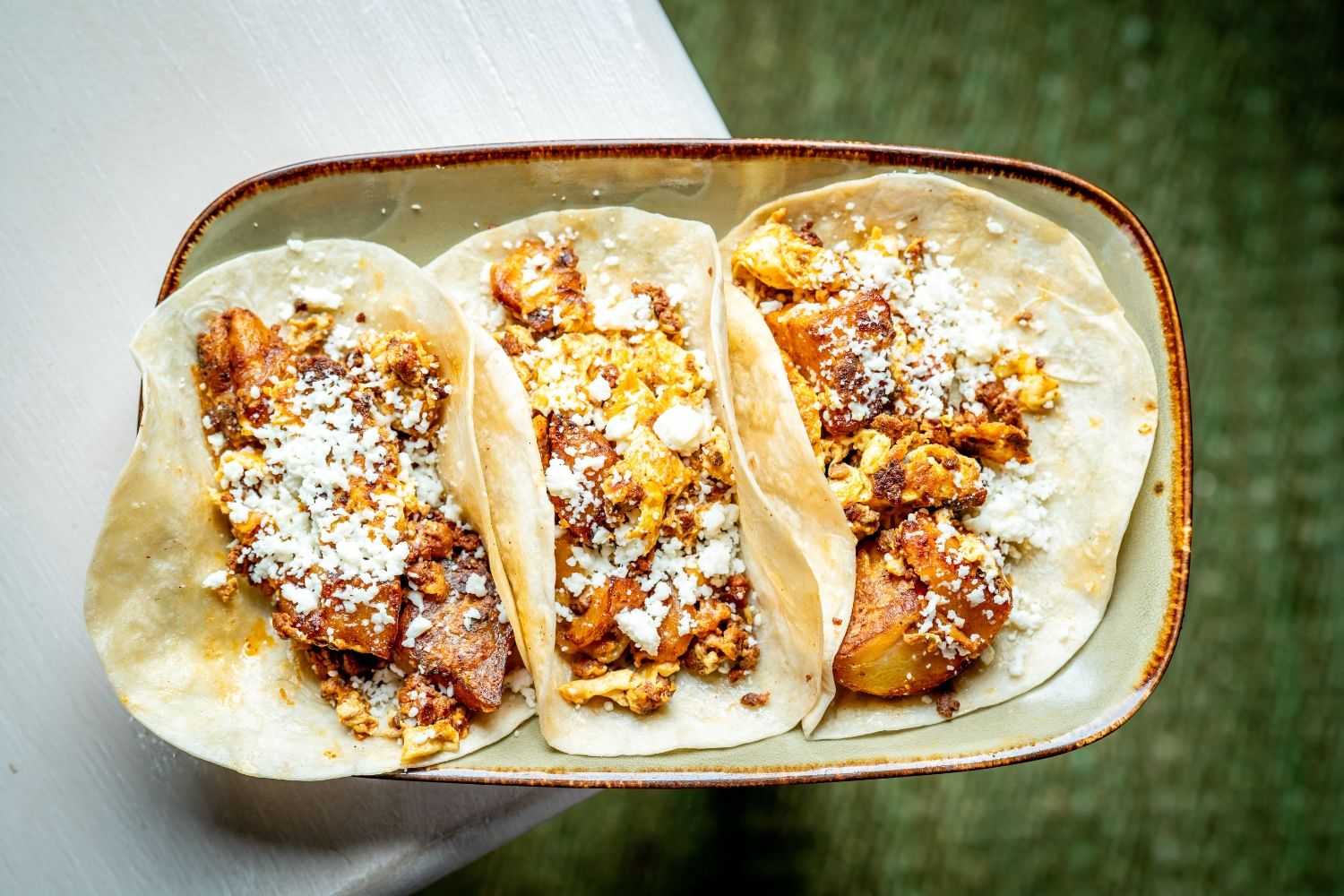 Three tacos with savory meat, scrambled eggs, and cheese on a rectangular plate.